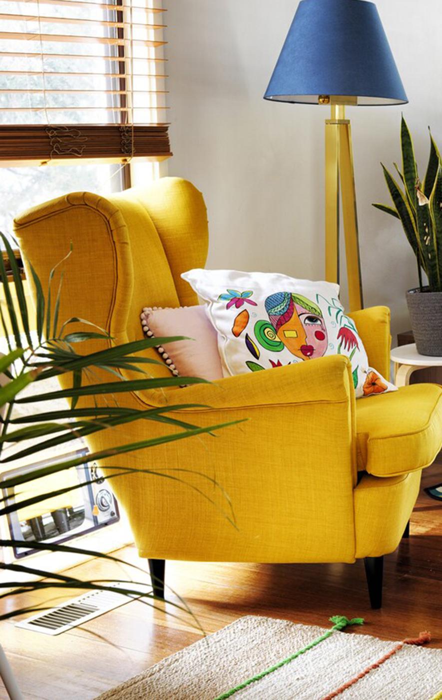 Séjour avec un fauteuil à oreilles jaune, coussin brodé multicolore et un lampadaire à abat-jour bleu foncé.