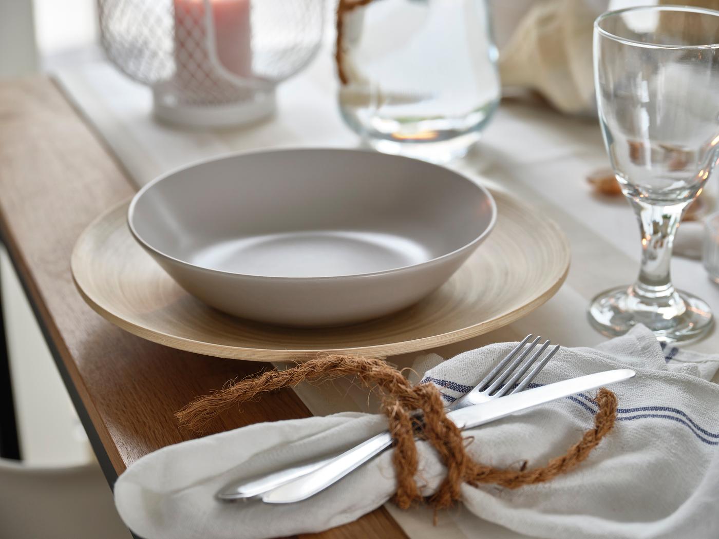 Servizio da tavola leggero composto da una ciotola su un sottopiatto in legno, un bicchiere, una candela da tè e un tovagliolo con posate.