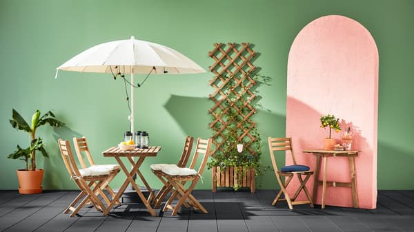 Set de muebles de jardín ASKHOLMEN color madera, incluidas mesas, sillas plegables y una jardinera. El fondo del espacio es verde y el piso es gris oscuro. 
