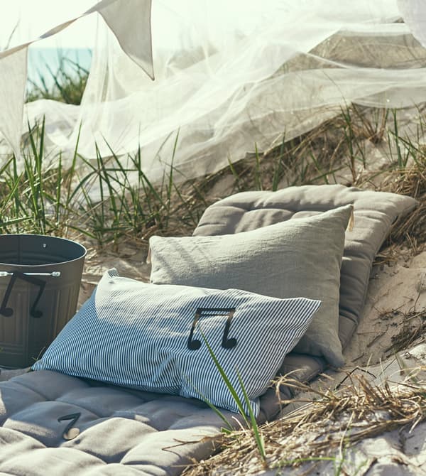 Several cushions sit in the sand next to a DIY speaker made from a bucket.