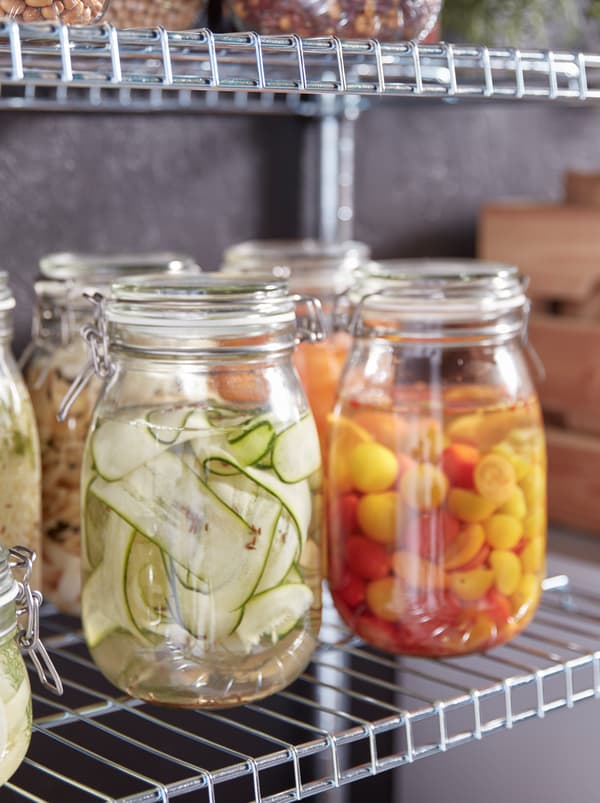 Several KORKEN glass jars with lids filled with colourful pickled vegetables of different kinds, standing on metal shelves.