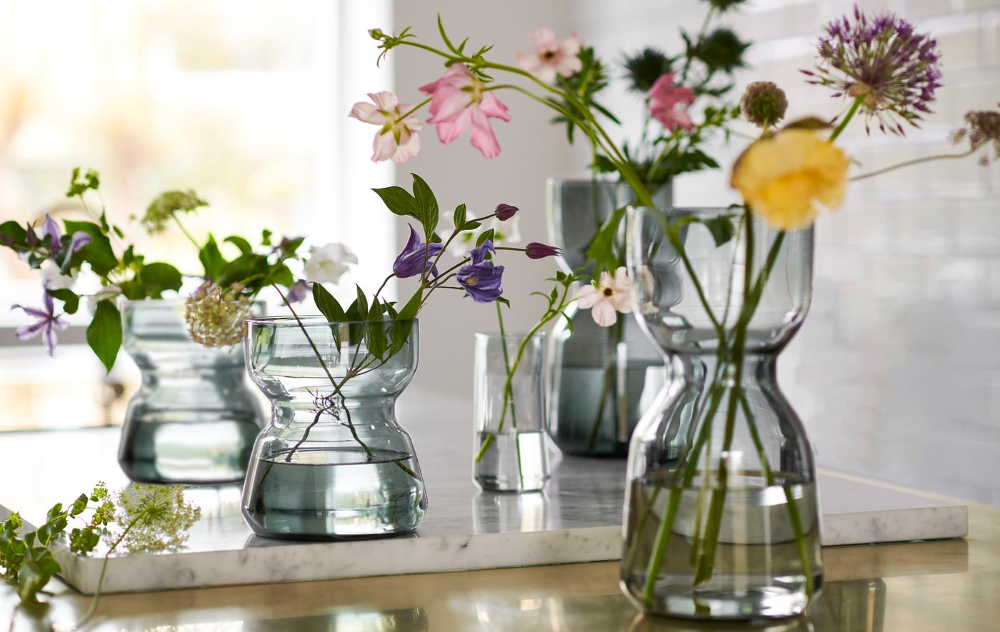 Several OMTÄNKSAM glass vases on a counter, with characteristic waists to facilitate grip, fresh flowers in each.
