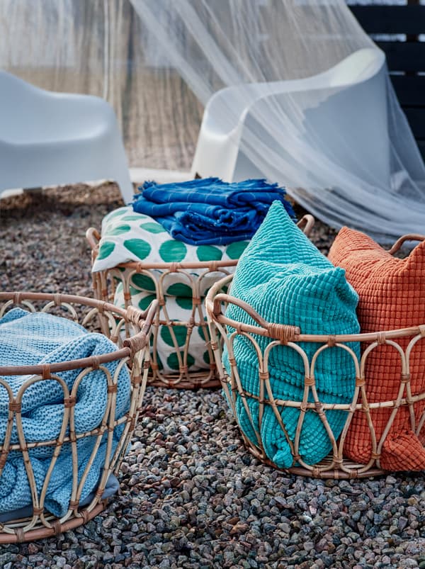 Several SNIDAD rattan baskets standing on gravel, filled with INGABRITTA throws, GULLKLOCKA cushions and other textiles.