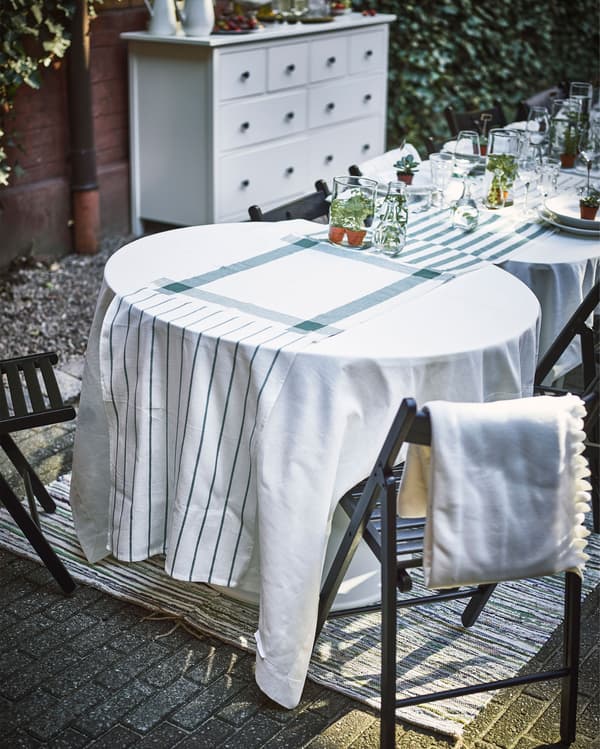 Several tables of different shapes and sizes sit in a row, with white tablecloths and a table runner made of white and green dish towels.