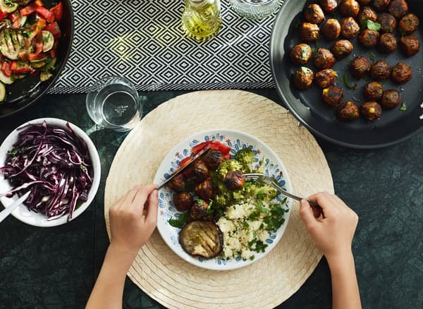 شخص يحمل سكينًا وشوكةً فوق طبق من الطعام على طاولة مع أطباق تقديم عليها خضروات وكرات خضروات.
