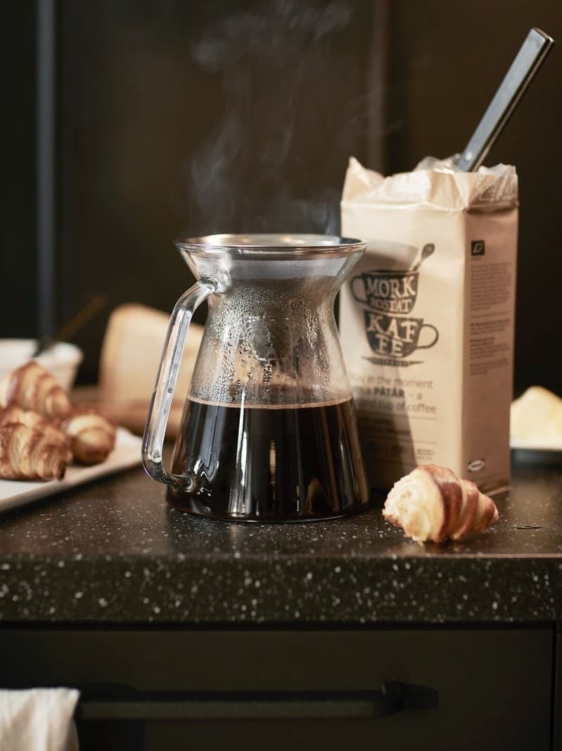 Skleněná konvice s čerstvě uvařenou kávou IKEA na černé pracovní desce, vedle otevřený sáček mleté kávy IKEA MÖRK ROST a croissanty na talíři.