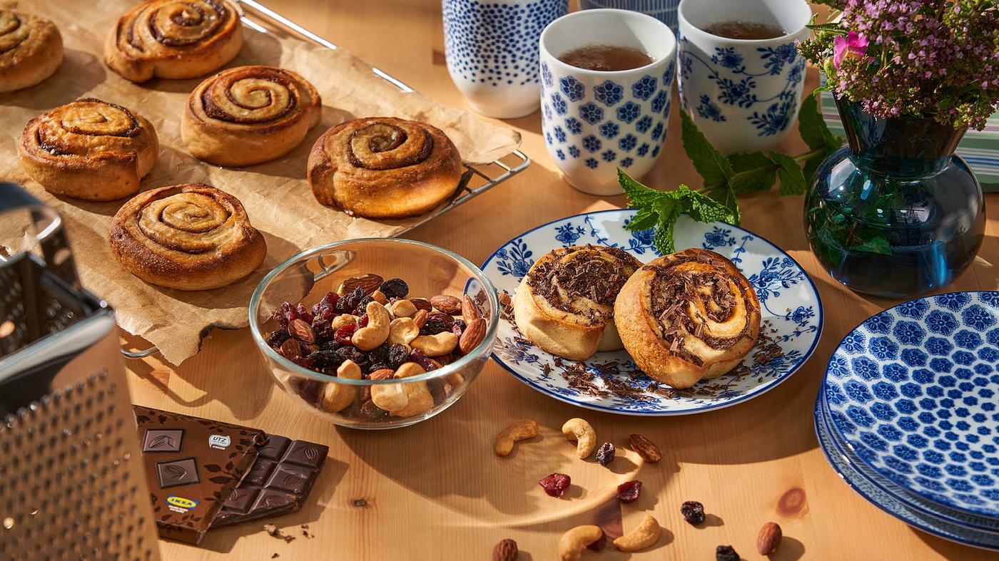 Skořicové rolky KAFFEREP na chladicí mřížce LÄTTBAKAD, před nimi miska BLANDA s oříšky MUNSBIT, dezertní talíře ENTUSIASM se dvěma rolkami s nastrouhanou čokoládou, hrnky ENTUSIASM s čajem a váza/svícen STOCKHOLM 2017.