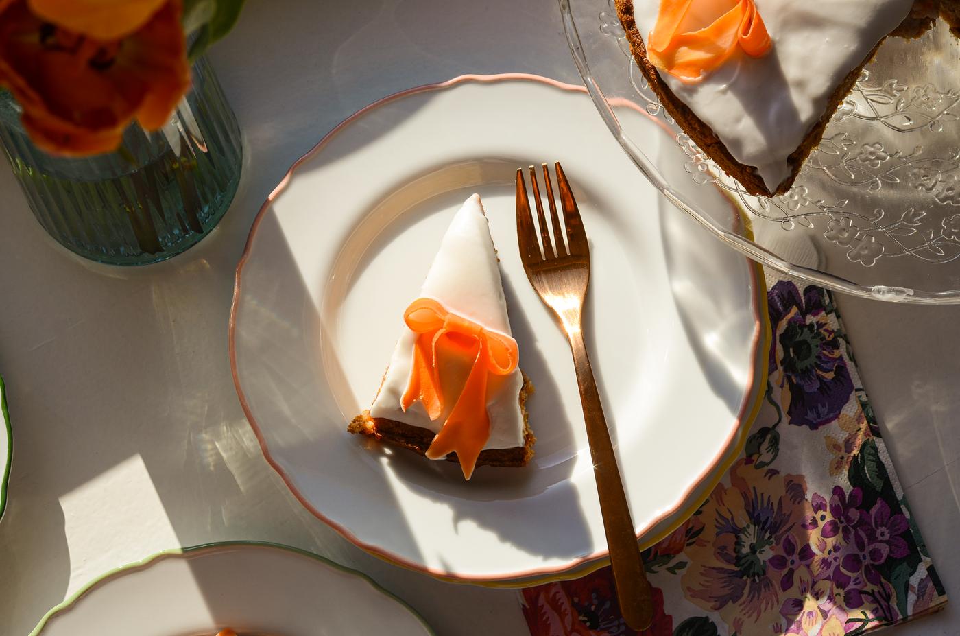 Slice of iced cake with orange flower decoration on white plate next to gold fork, with a vase of orange flowers nearby.
