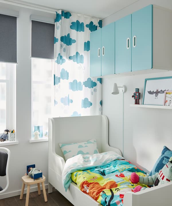 Small extendable bed in corner of a child’s room. Colourful bedlinen and wall cabinets. Window with curtain and roller blind.