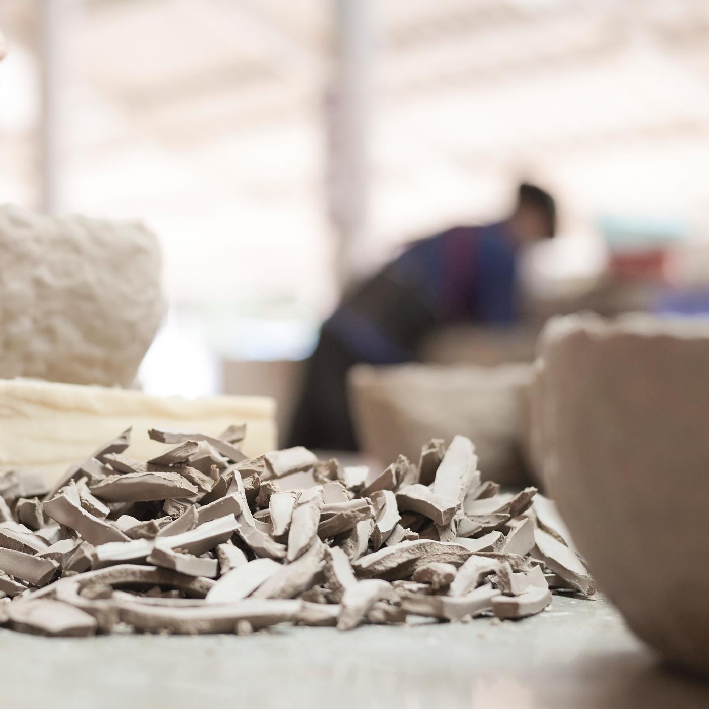 Small pieces of broken pottery on a table with larger intact ceramics and an artisan in the background.