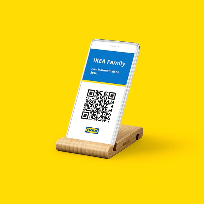 Smartphone displayed on a wooden IKEA stand, showing an IKEA Family digital membership card with a QR code, Lithuanian email address, and the word 'Lietuva', set against a bright yellow background.