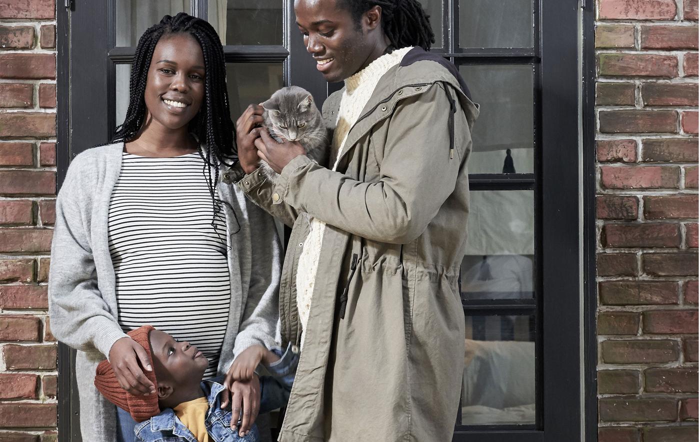 Smiling, late-twenties man and pregnant woman in front of a brick wall, a small boy in the middle; the man holds a cat.