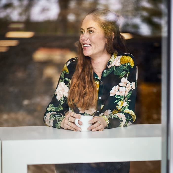 Smiling woman with long red hair sitting at a white table, holding a white mug, and looking out the window. She is wearing a dark floral blouse with green, yellow, and white patterns.