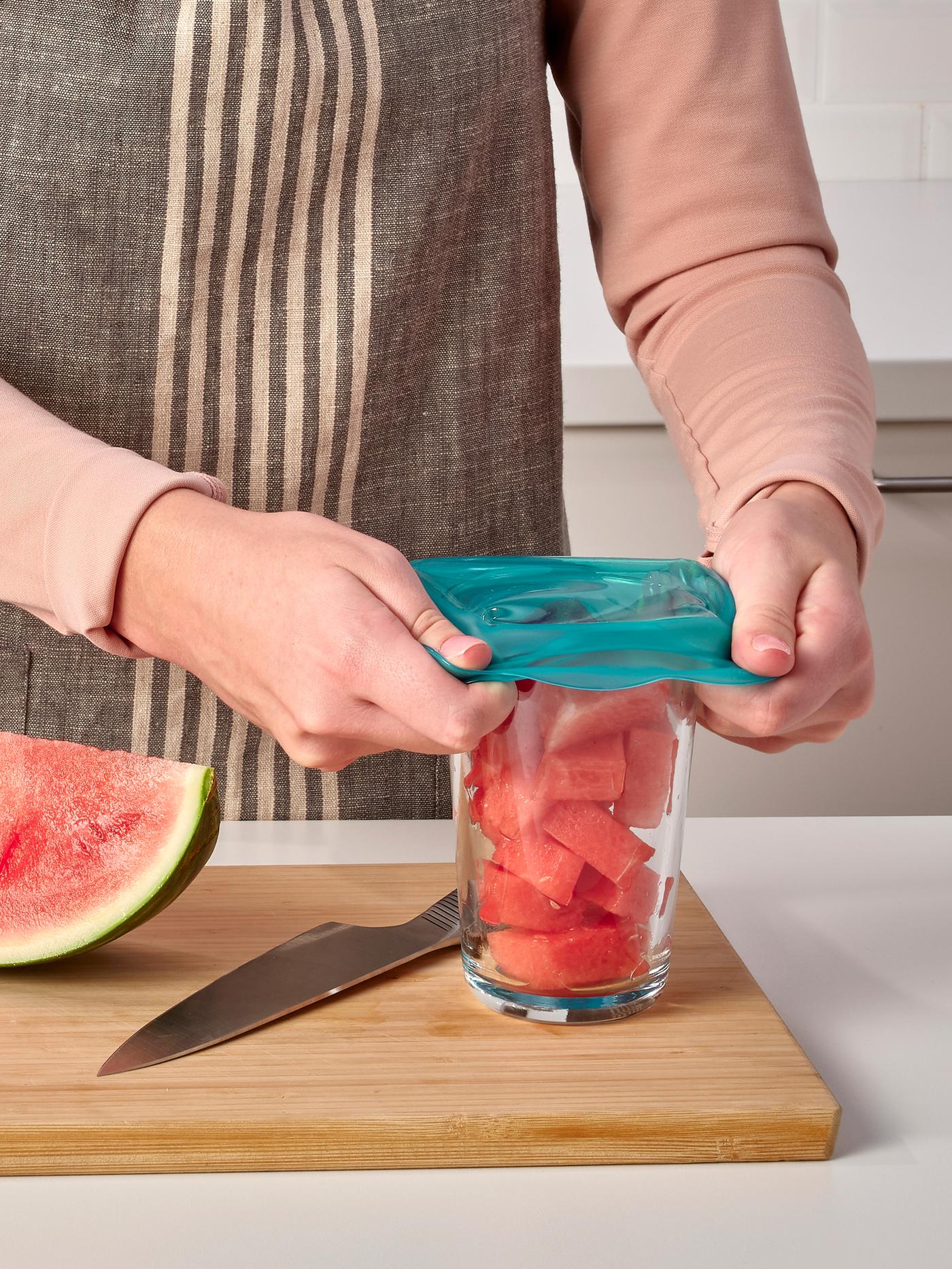 Sobre una tabla para picar de madera, una persona cubre un vaso de sandía picada con una cubierta de silicón reutilizable ÖVERMÄTT para alimentos.