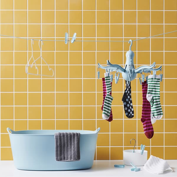 Socks on a blue PRESSA hanging dryer, pegs and hangers on a clothes line above a blue TORKIS laundry basket against a yellow tiled wall.
