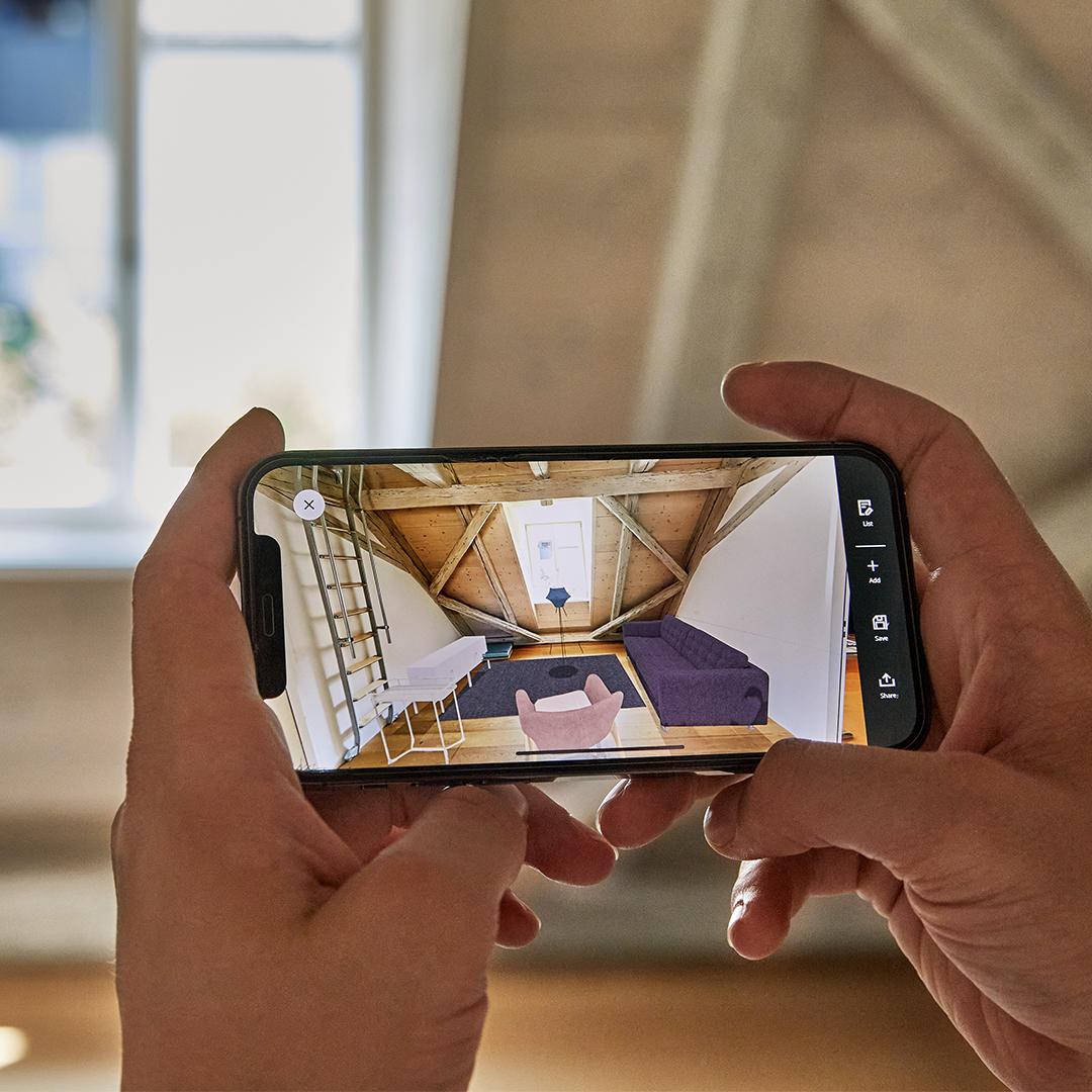 Someone holds their smartphone horizontally to design a room in mixed reality using the IKEA Kreativ 3D room planner.