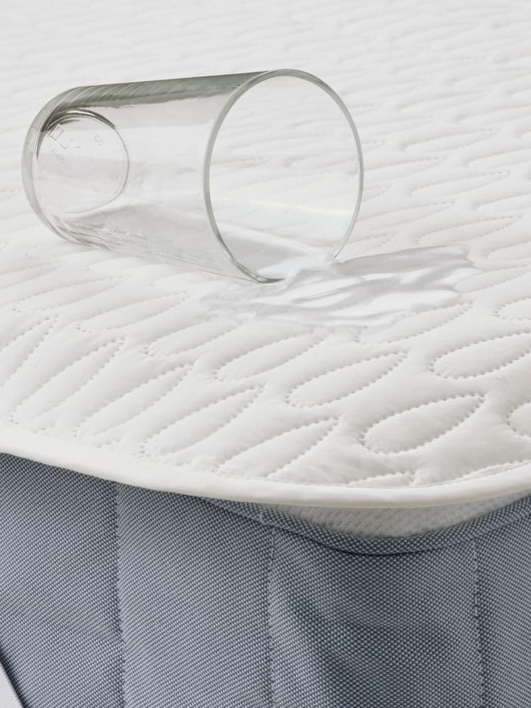 SOTNÄTFJÄRIL mattress protector is laid on a mattress with a glass of water being spilled on the mattress, showing its waterproof protection.