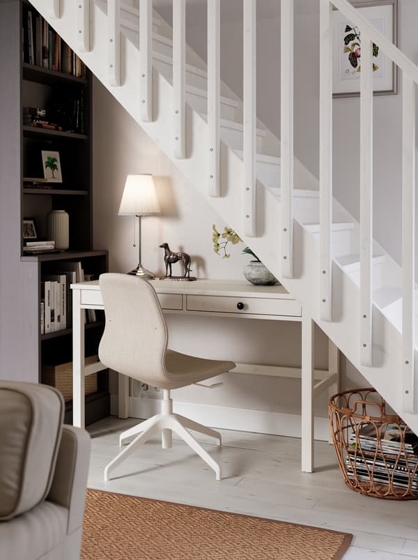 Sous les escaliers, un espace de travail composé d’un bureau HEMNES blanc, d’une chaise LÅNGFJÄLL, d’une lampe et d’une grande bibliothèque.