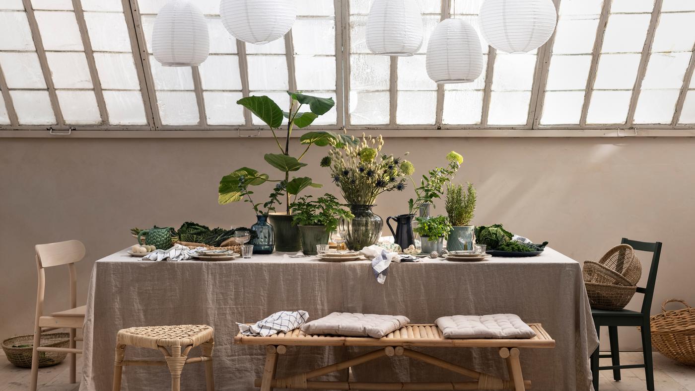 Sous un toit de verre, des assiettes, des plantes et des feuillages dans des cache-pots, ainsi que des vases sont disposés sur une table munie d’une nappe en lin.