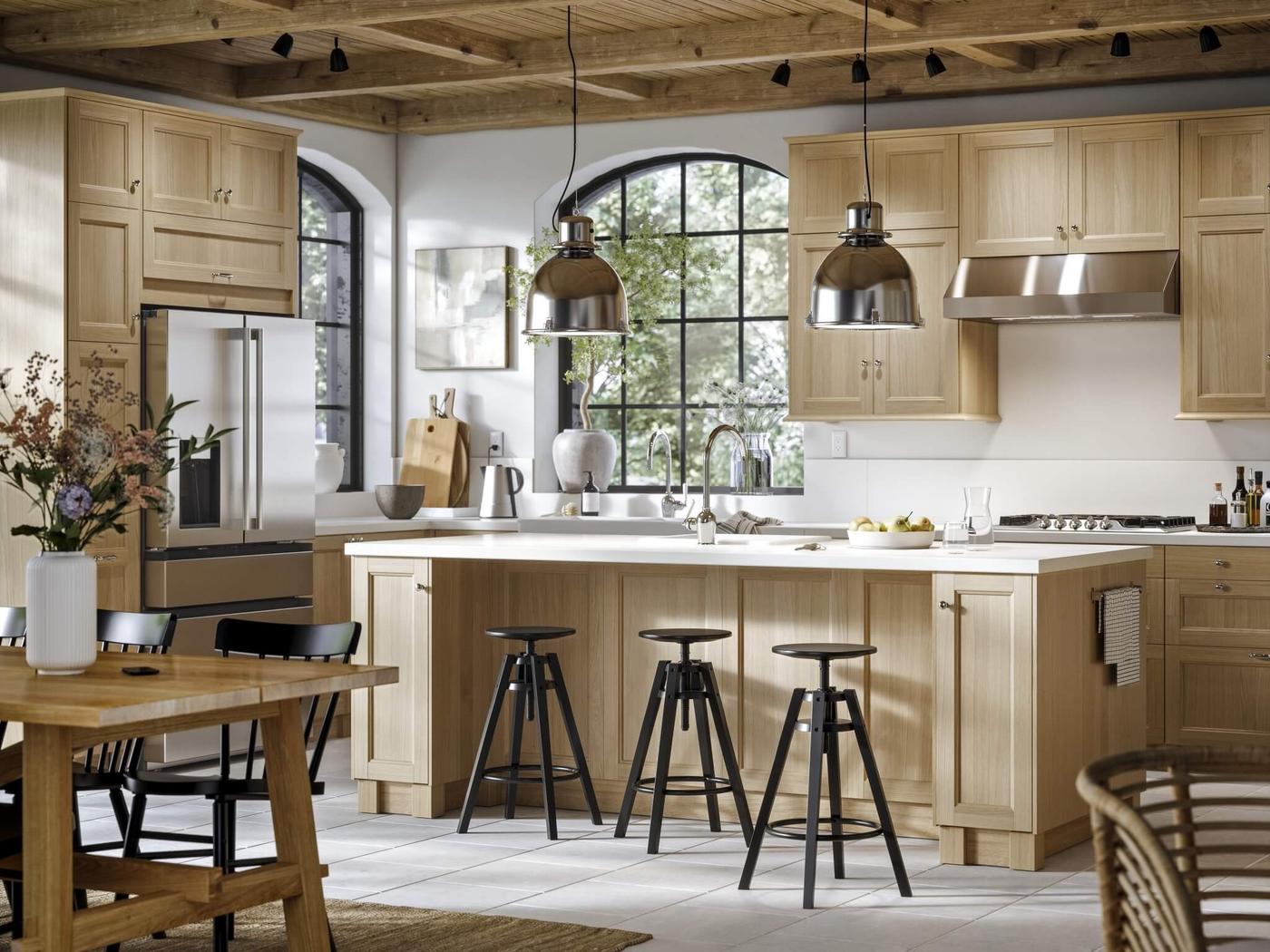 Spacious and well-lit modern kitchen with wooden cabinets, a central island with bar stools, stainless steel appliances, decorative pendant lighting, and a small wooden table in the background decorated with a vase of flowers.