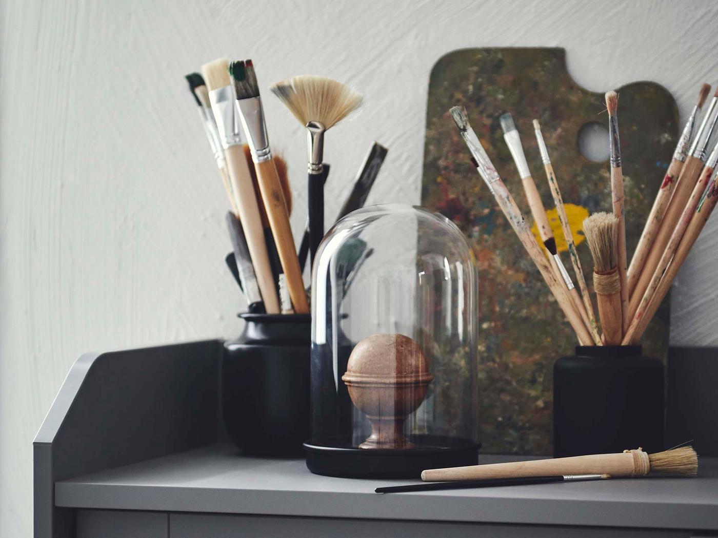 Sprawling bouquets of thin paintbrushes placed in a black KONSTFULL vase and a black jar atop a HAUGA cabinet.