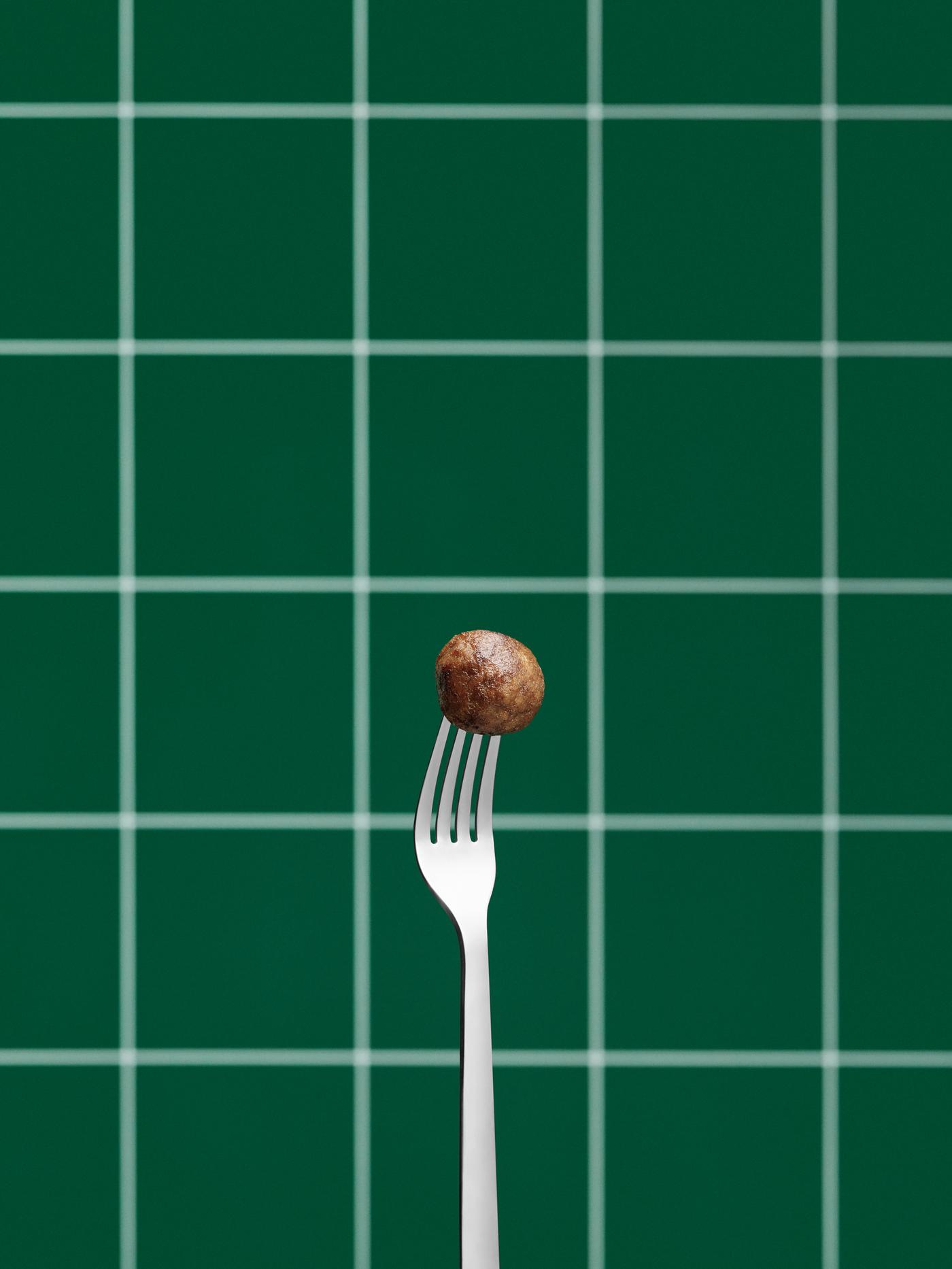 Stainless steel fork held upright, its tines stuck into a single plant-based meatball. Green, tiled wall in the background.