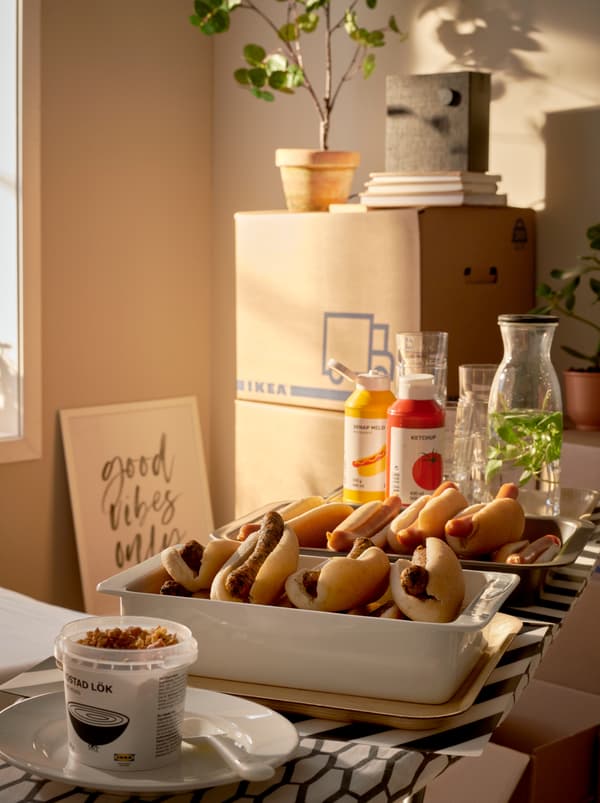 Stanza soleggiata con cartoni da imballaggio JÄTTENE impilati e un buffet servito su un’asse da stiro DÄNKA, con teglie di hot dog appoggiate su dei vassoi - IKEA