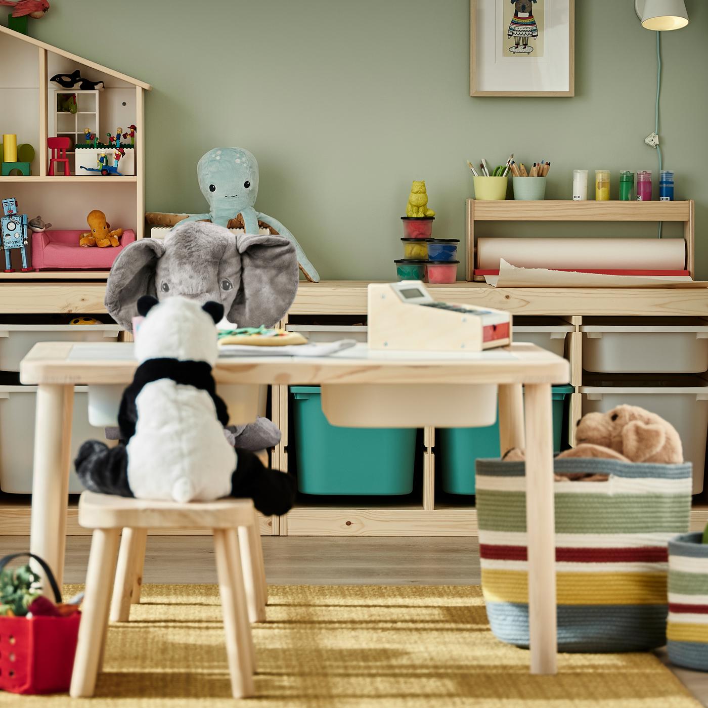 Stofftiere sitzen rund um einen Kindertisch mit Hockern. Im Hintergrund sind Gestelle mit Spielzeugaufbewahrungen in einem Kinderzimmer zu sehen.