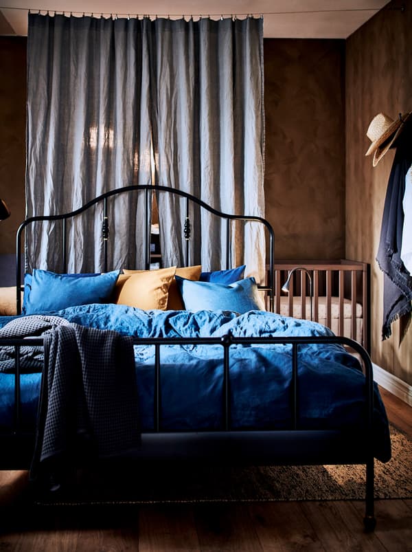 Structure de lit SAGSTUA en métal noir au milieu d’une chambre à coucher divisée par des rideaux et dans laquelle on trouve aussi un lit d’enfant.