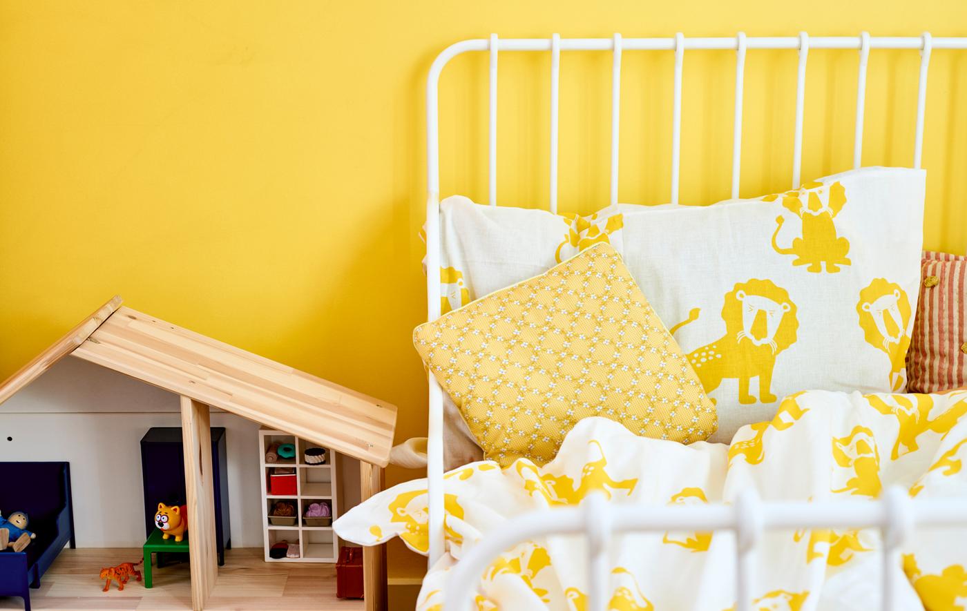Struttura letto bianca, federe e copriletto bianchi con leoni gialli e una casa delle bambole in legno sullo sfondo di una parete gialla - IKEA
