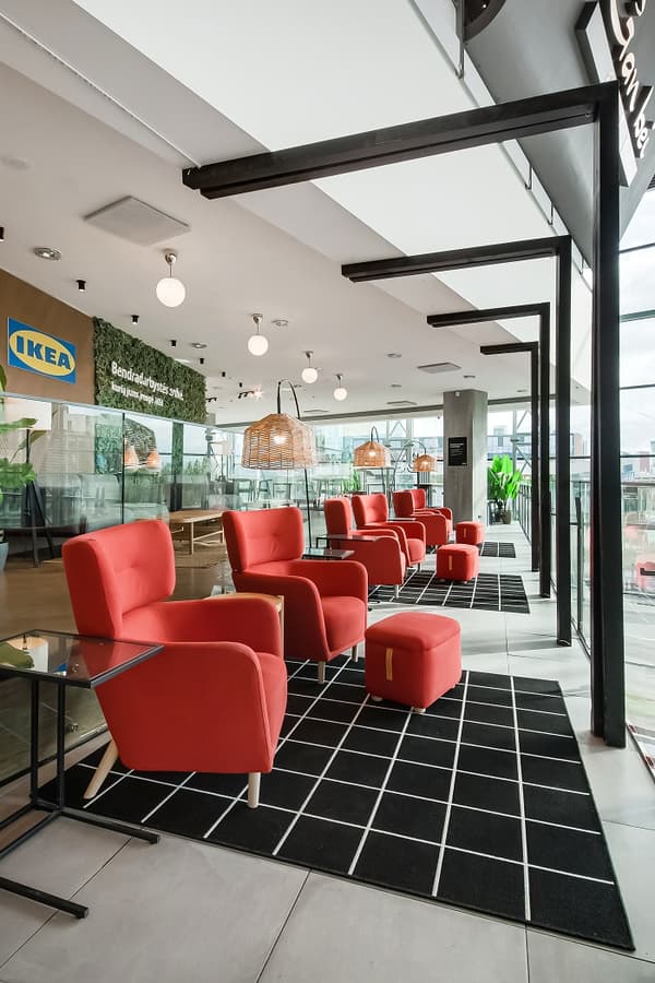 Stylish IKEA lounge area with a row of bright red armchairs and matching ottomans on black grid-patterned rugs, wicker pendant lamps, floor-to-ceiling windows, and modern lighting in a spacious showroom.
