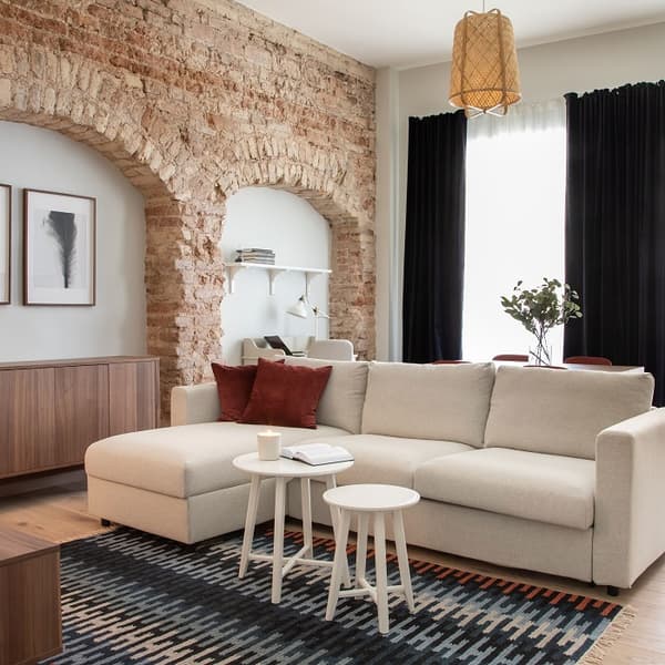 Stylish living room blending rustic and modern elements, featuring an exposed brick arch wall that separates the lounge from a study nook. A light beige sectional sofa with red velvet accent pillows sits atop a geometric-patterned rug. In front of the sofa are two small white nesting tables, one holding a candle and an open book. The room is framed by floor-length black curtains that contrast with the white walls and natural wood flooring. A wooden sideboard and a woven pendant light add warmth, while minimalist artwork and a vase with greenery complete the cozy, contemporary look.