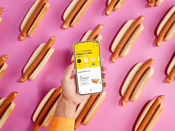 Su uno sfondo grafico con diversi hot dog IKEA, una mano in primo piano tiene uno smartphone sul quale è visualizzato un pasto ordinato nell’app IKEA.