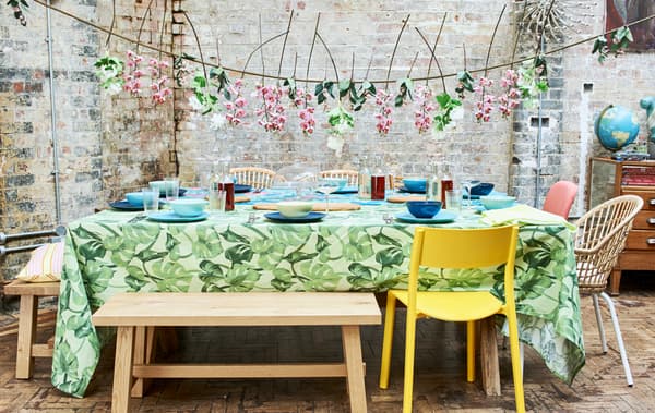 Summer party table with bright tablecloths, blue and green crockery set out, chairs and benches and a floral hanging garland.