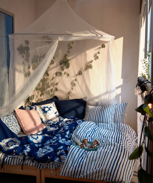 Sunlit balcony with double bed softly piled with cushions and blue-white bedlinen; mosquito net above, plants around.