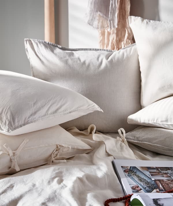Sunlit bedscape laden with pillows and cushions in light-coloured textiles, an open book lying on the duvet.