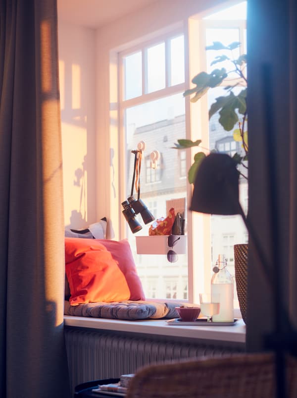 Sunlit window with a cityscape backdrop. On the sill a VIPPÄRT chair cushion, a tray of refreshments, and a large plant.
