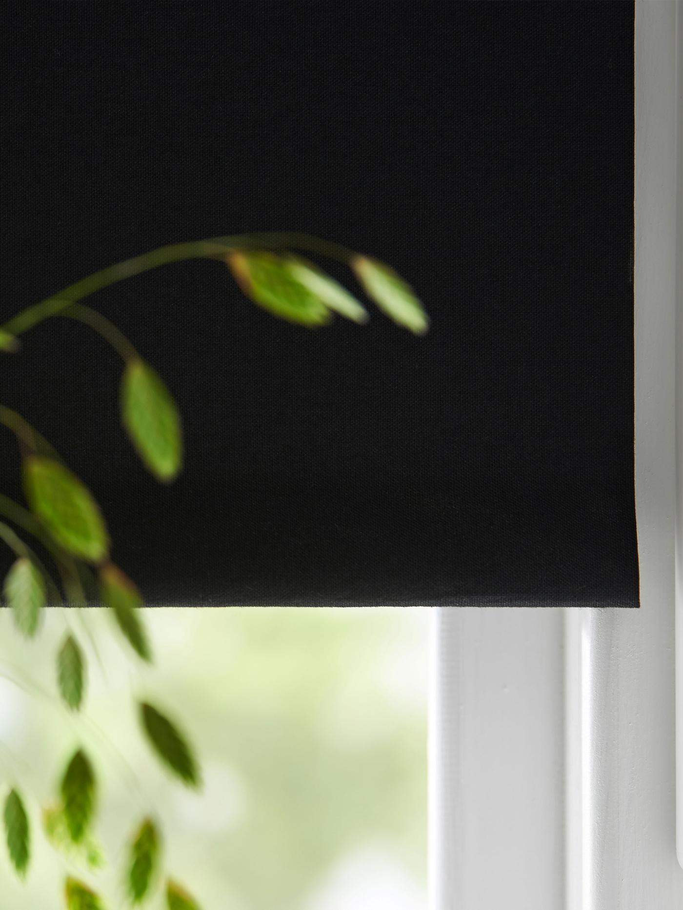 Svart mörkläggande rullgardin neddragen framför ett fönster, med gröna blad synliga vid sidan.