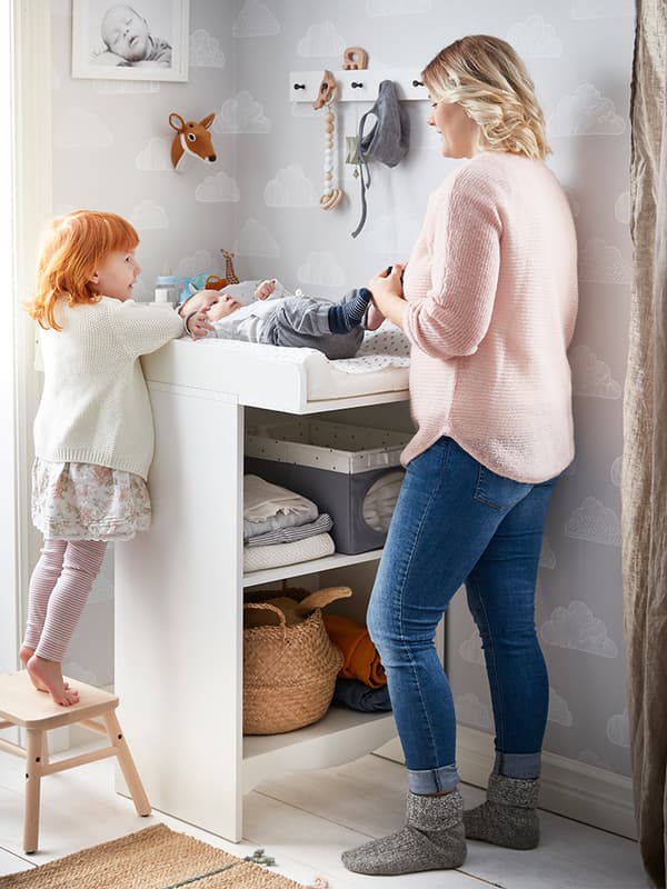 Table à langer SMÅGÖRA dans une chambre aux murs ornés de papier peint fleuri et où se trouvent armoire-penderie, fauteuil, mobile et articles de soins pour bébé.
