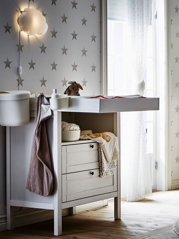 Table à langer SUNDVIK blanche, vêtements et draps pour bébé, lit à barreaux et petite table d’appoint.