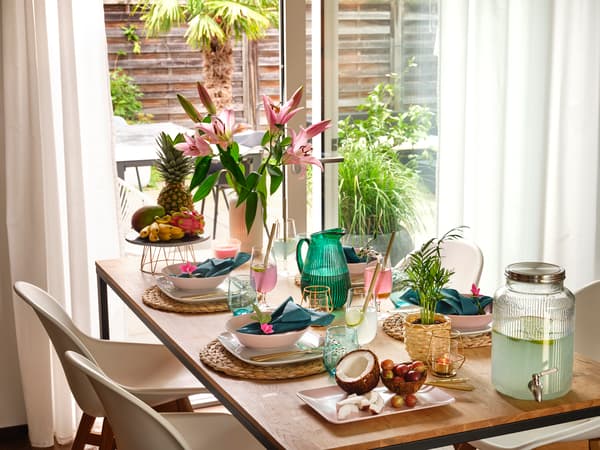 Table à manger d'inspiration tropicale, décorée de lys, d'ananas, de noix de coco, de verres et d'un distributeur de boissons près de la fenêtre.