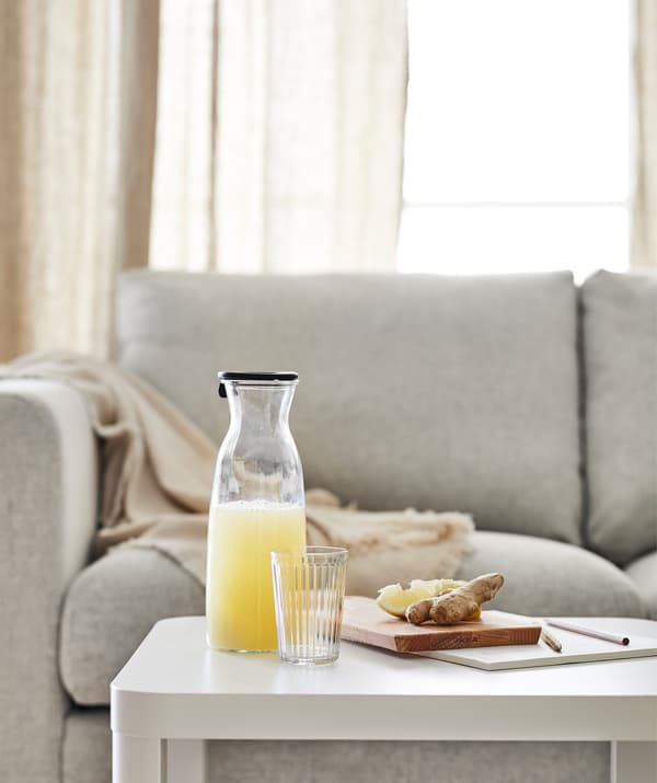 Table d’appoint TINGBY devant un canapé, surmontée d’une boisson fruitée dans une carafe et d’une planche à découper avec des morceaux de gingembre et de citron dessus.