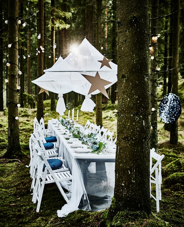 Table de noces dans la forêt avec longue table blanche, chaises pliantes blanches, objets décoratifs suspendus et guirlandes lumineuses enroulées autour d’arbres