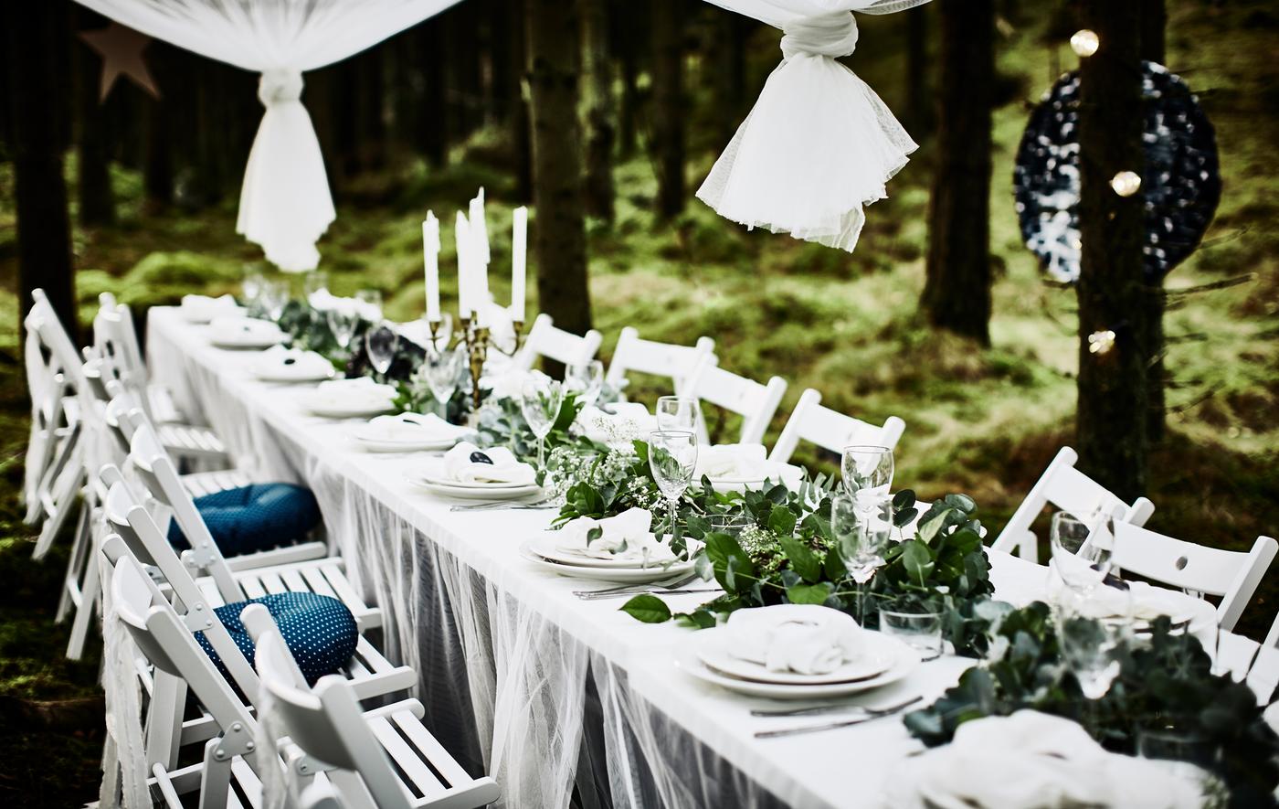 Table de noces en forêt, avec plateau de table blanc, rideaux fins comme nappe, chaises pliantes blanches, vaisselle blanche et chemin de table central réalisé avec de la verdure