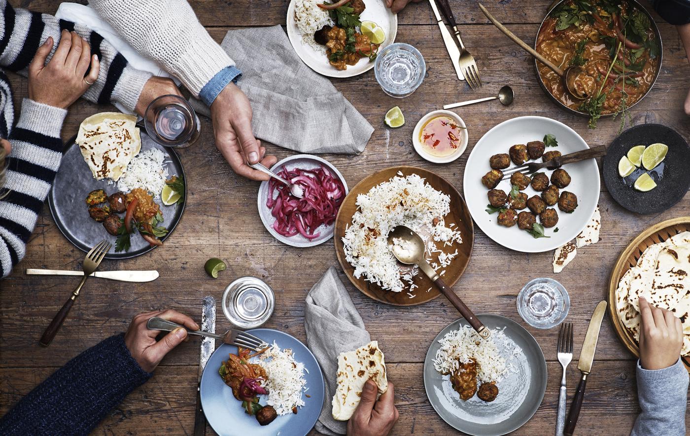 Table en bois sur laquelle sont présentés des boulettes, du riz et des légumes en saumure, avec couverts et mains qui se servent.