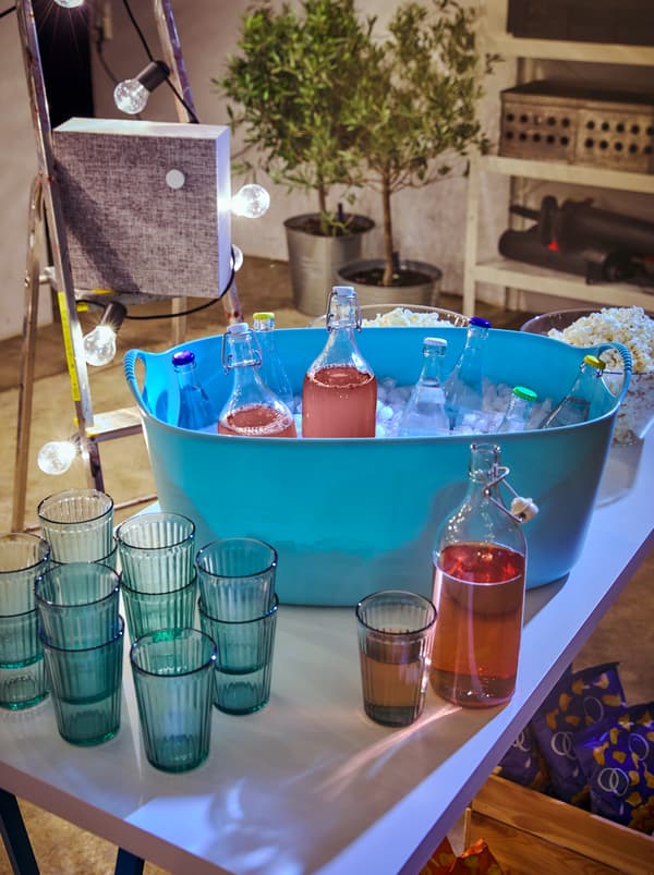 Table with stacked KALLNA glasses by a TISKEN laundry basket filled with ice and capped drinks, including KORKEN glass bottles.
