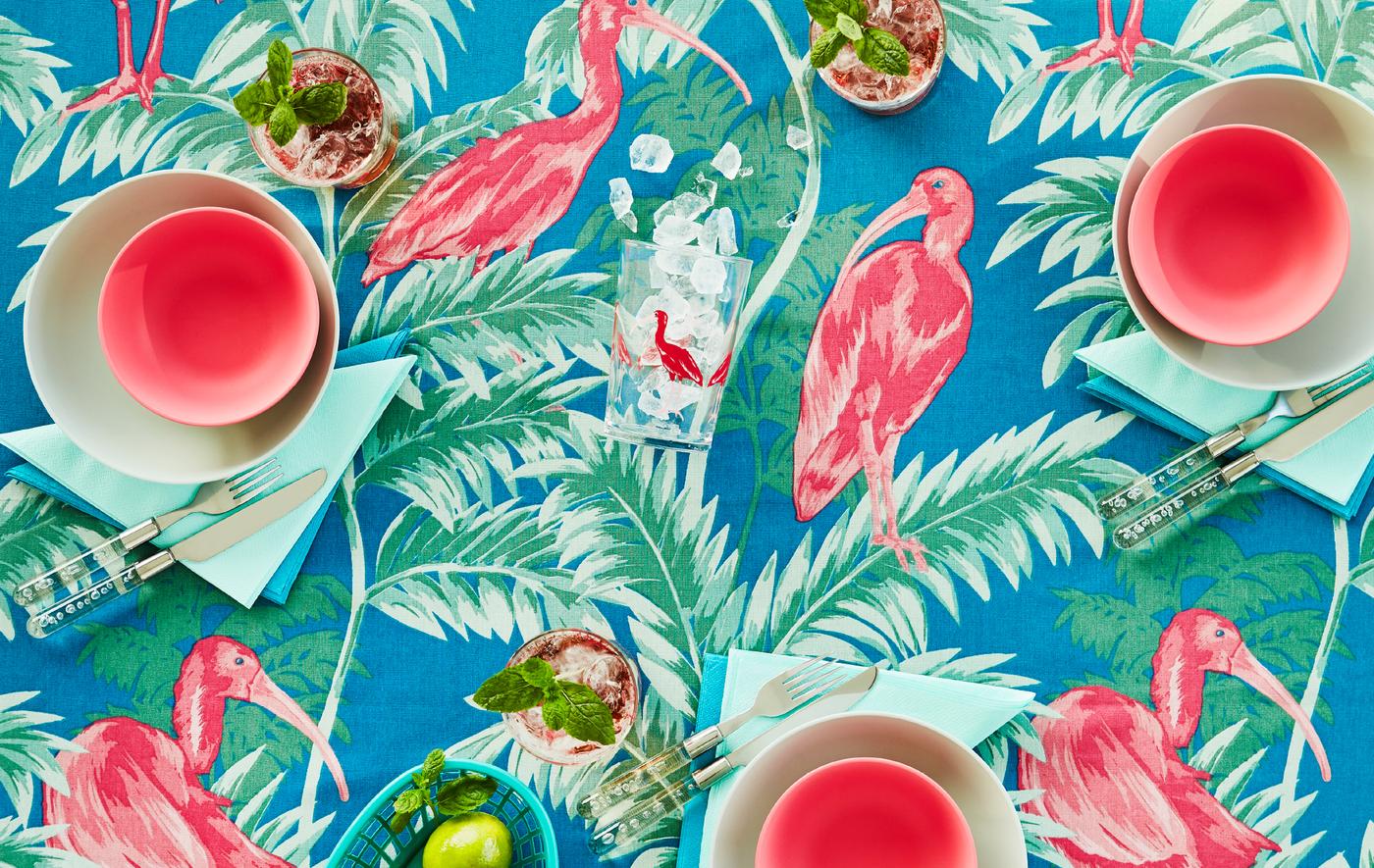Tabletop with a cloth with a colourful, tropical pattern in pink, green, and blue. The table is set with tableware to match.