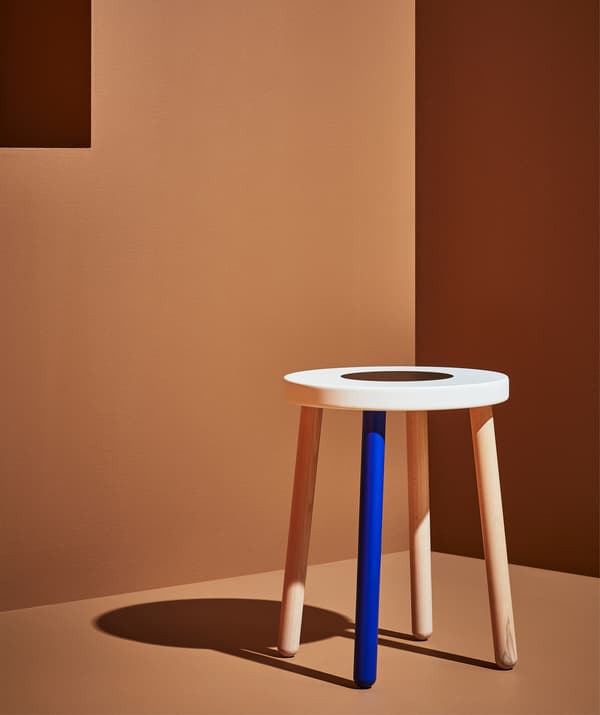 Tabouret en bois avec un pied bleu et un plateau blanc, dans une chambre orange.