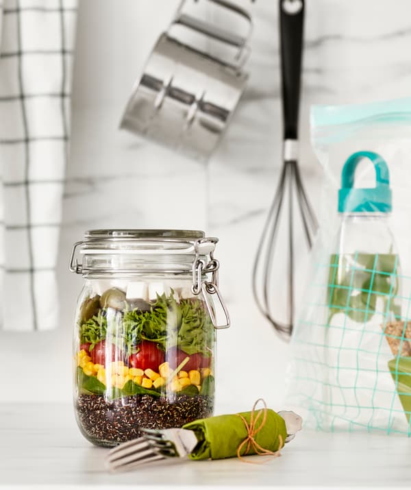 Tall glass jar with fastened lid, holding a layered, Greek-style salad; eating-utensil kit and water bottle on the side.