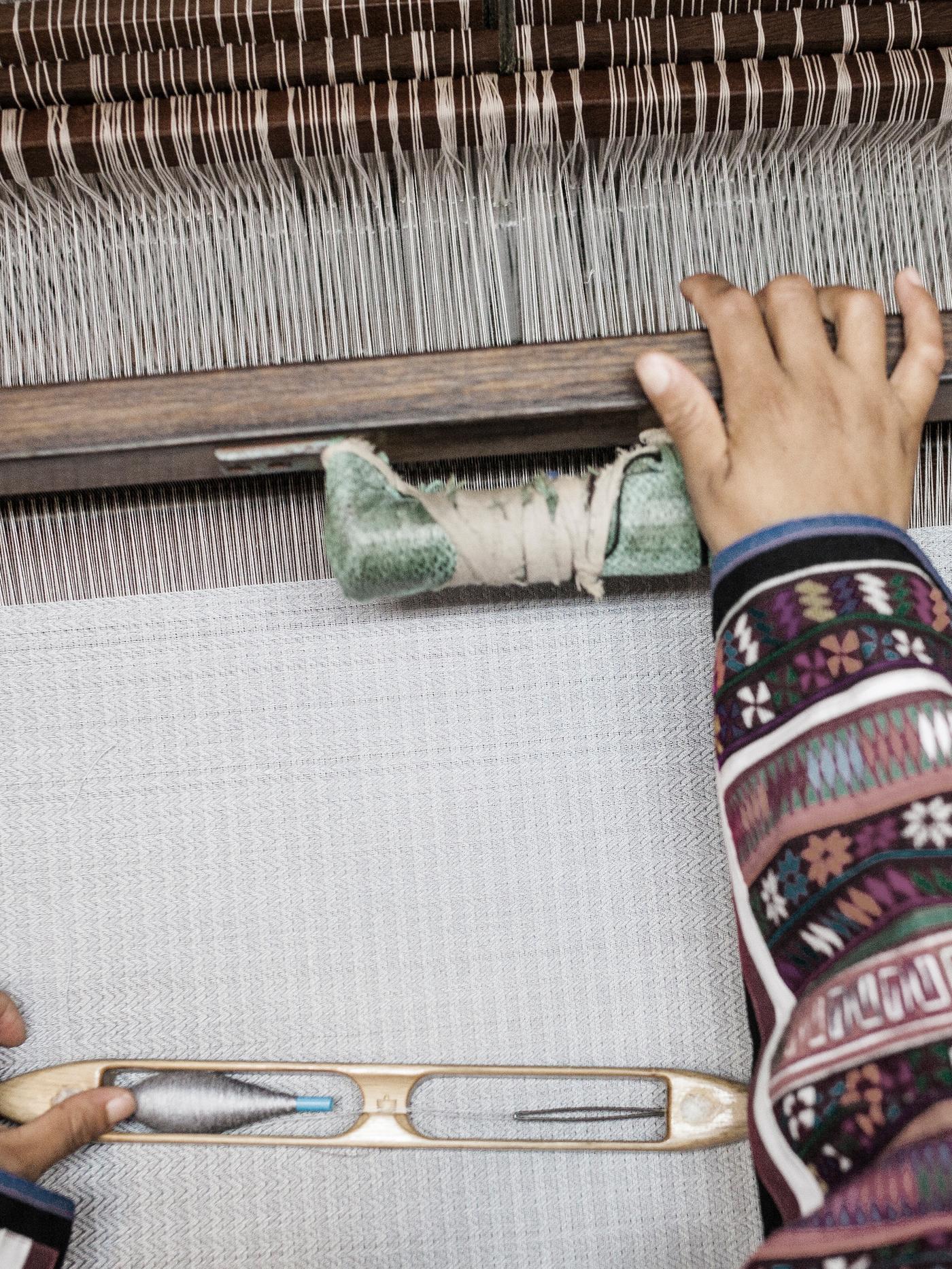 Tangan seorang penenun yang memakai baju bercorak tradisional sedang menenun ambal berwarna putih menggunakan alat tenun.